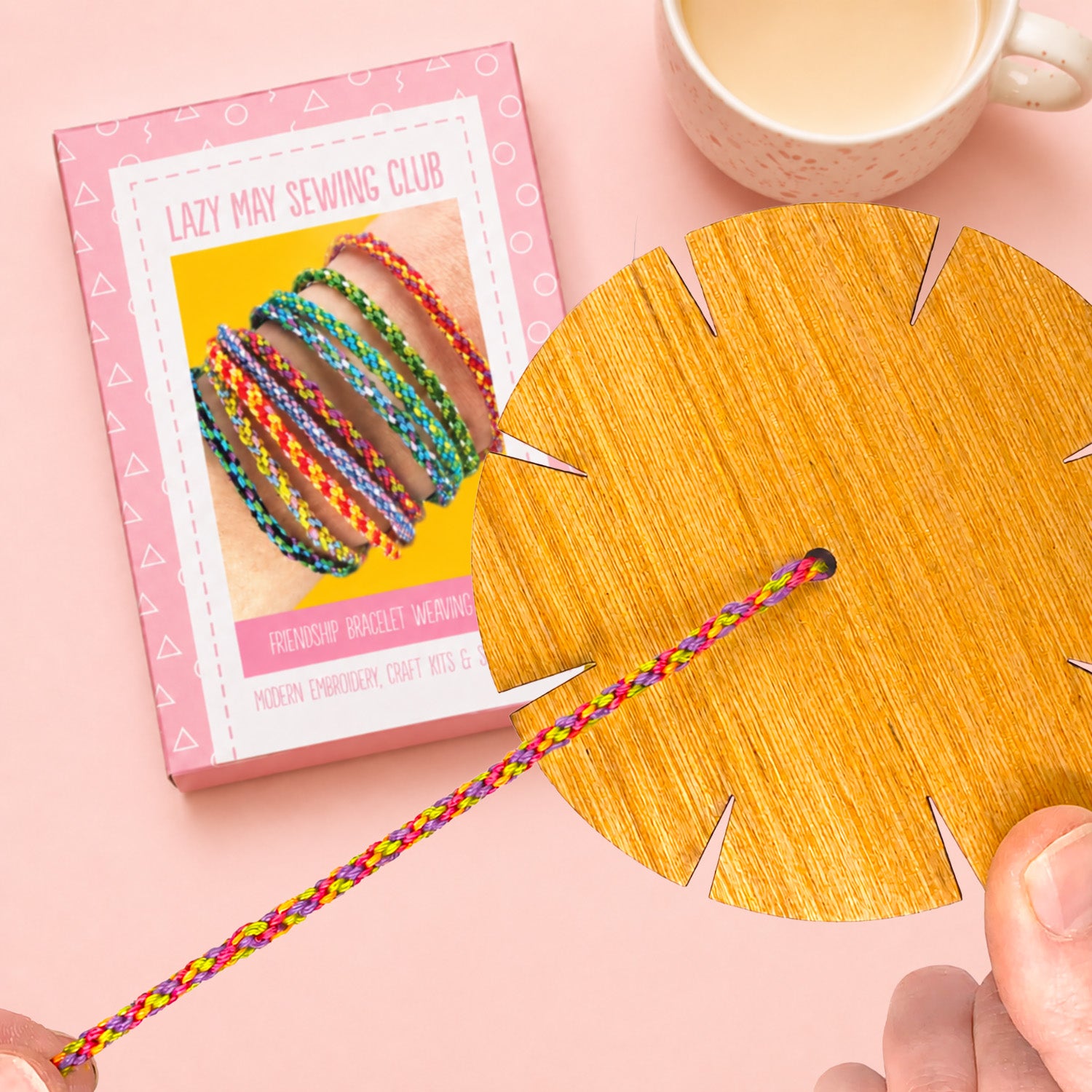 A craft kit box labeled 'Lazy May Sewing Club' sits on a pink background next to a large circular wooden disc with cutout sections and a braided bracelet threaded through the center. A hand is holding the bracelet, and a cup is partially visible at the top right.