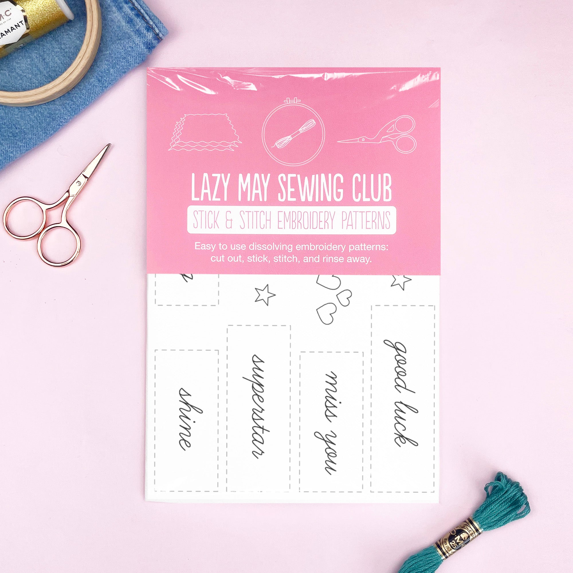 Embroidery pattern sheet with secret messages, surrounded by scissors, thread, and sewing tools on pink background