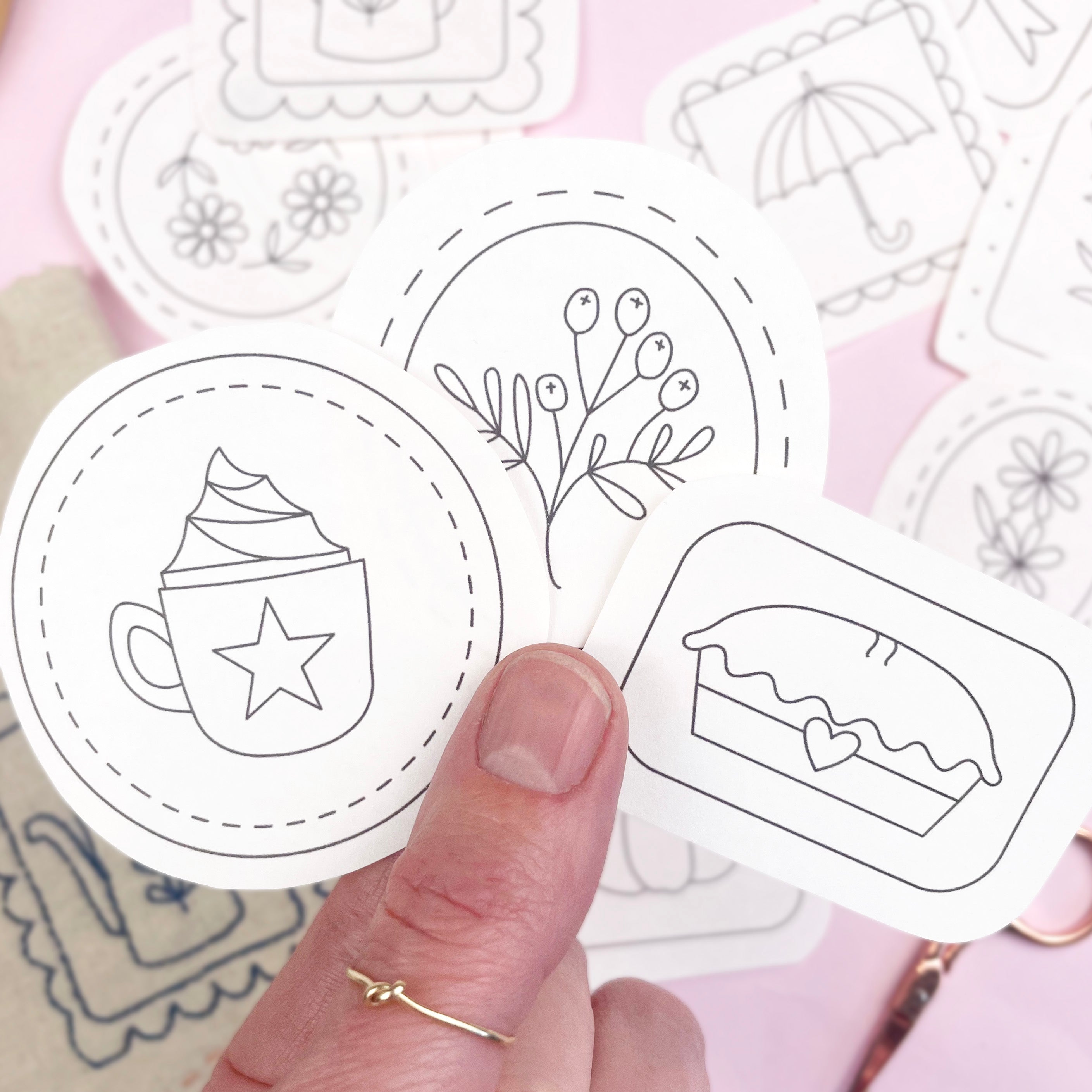 Hand holding a set of hand embroidery iron on transfers  on a pink background, the designs feature a hot chocolate, branches and a apple pie