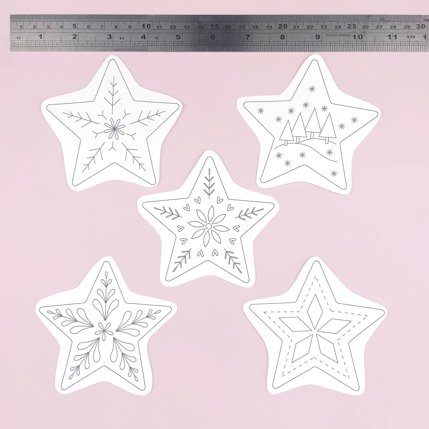 Five white star-shaped embroidery templates or patterns with black line-art holiday designs, arranged on a light pink background with a metal ruler at the top for scale.