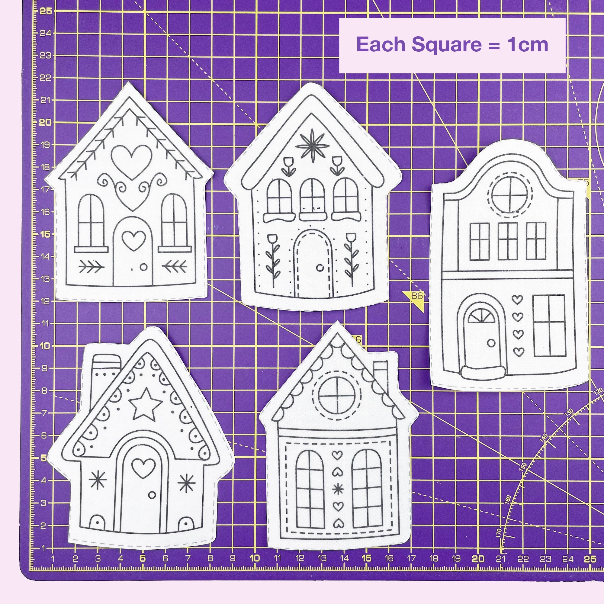 Five gingerbread house-shaped embroidery patterns on a purple cutting mat with grid lines.