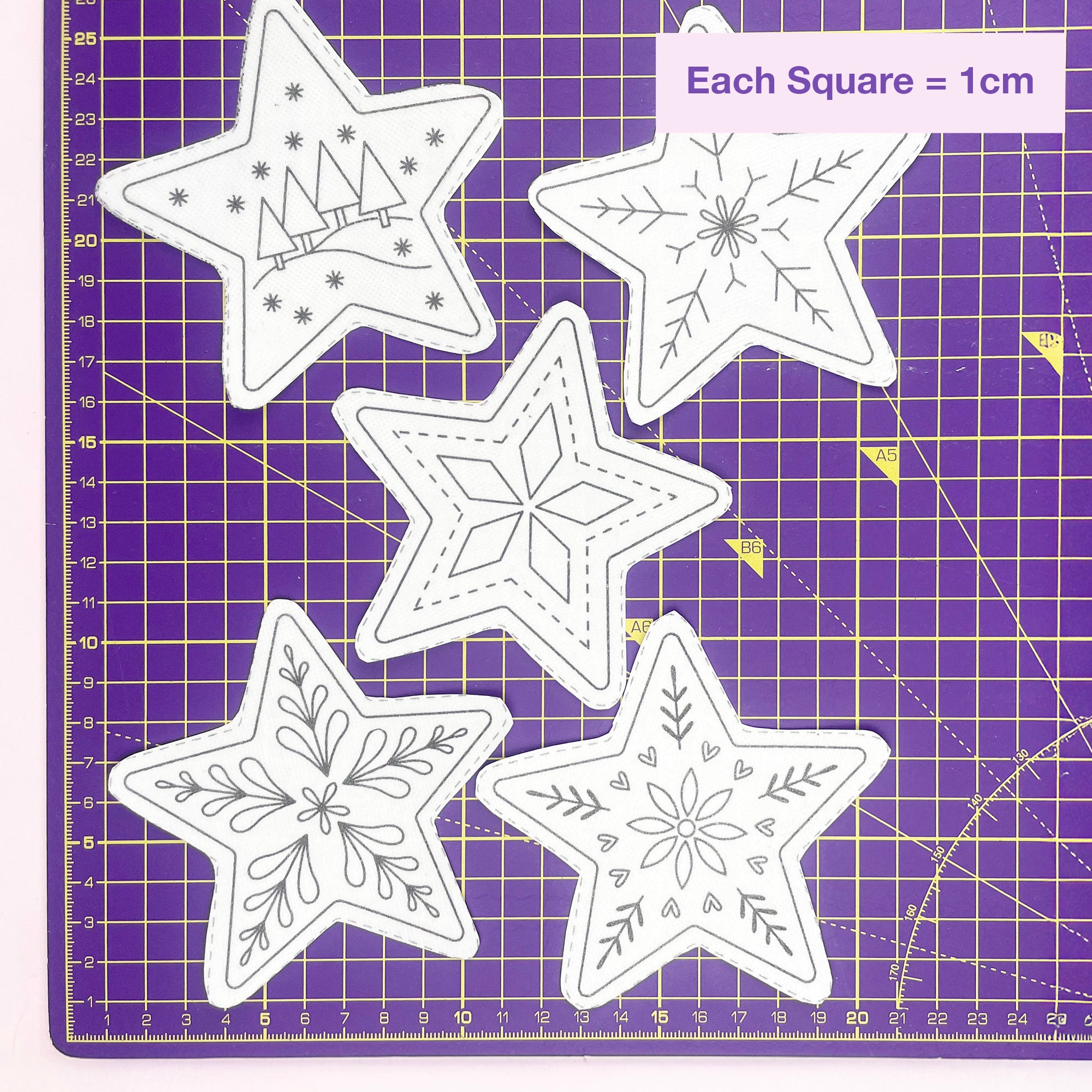 Five star-shaped embroidery patterns with different winter and floral line designs laid out on a purple cutting mat with a size reference label reading 'Each Square = 1cm'.