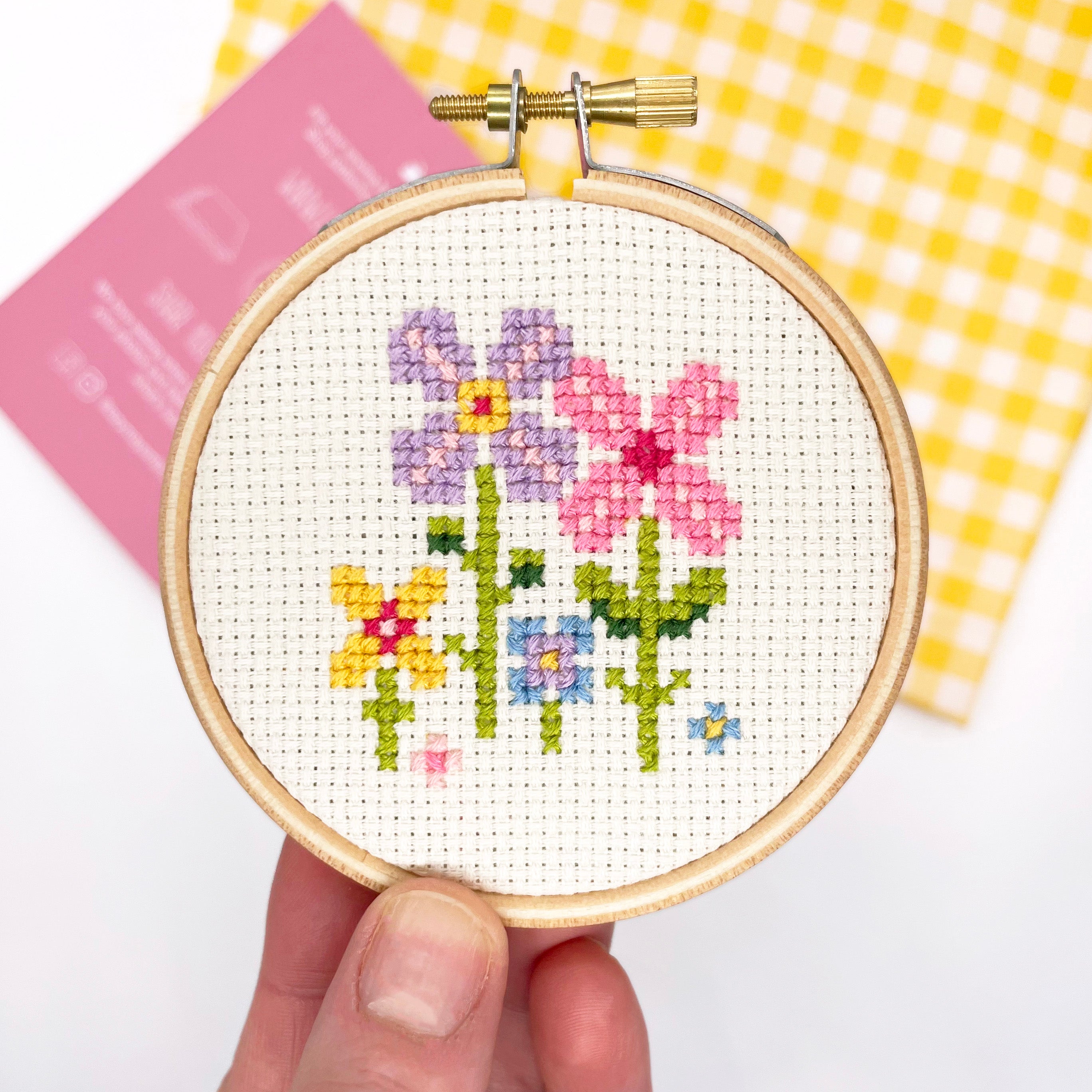 Hand holding a small floral cross stitch in a hoop from the Tiny Garden: Cross Stitch Kit with pink and yellow background