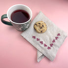 Decorative corner embroidery in pink stitched on a linen napkin, next to a cup of tea and a biscuit.