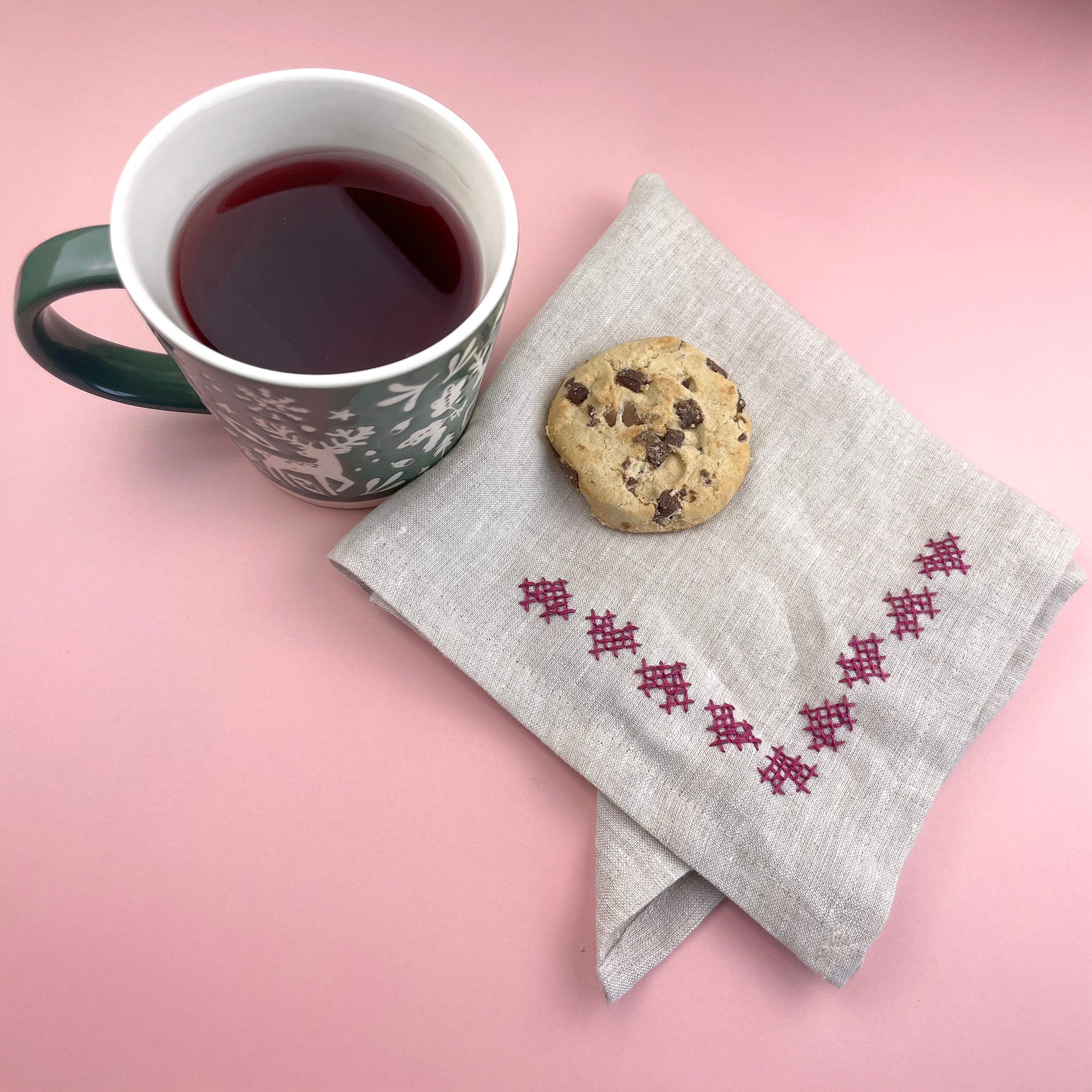 Decorative corner embroidery in pink stitched on a linen napkin, next to a cup of tea and a biscuit.