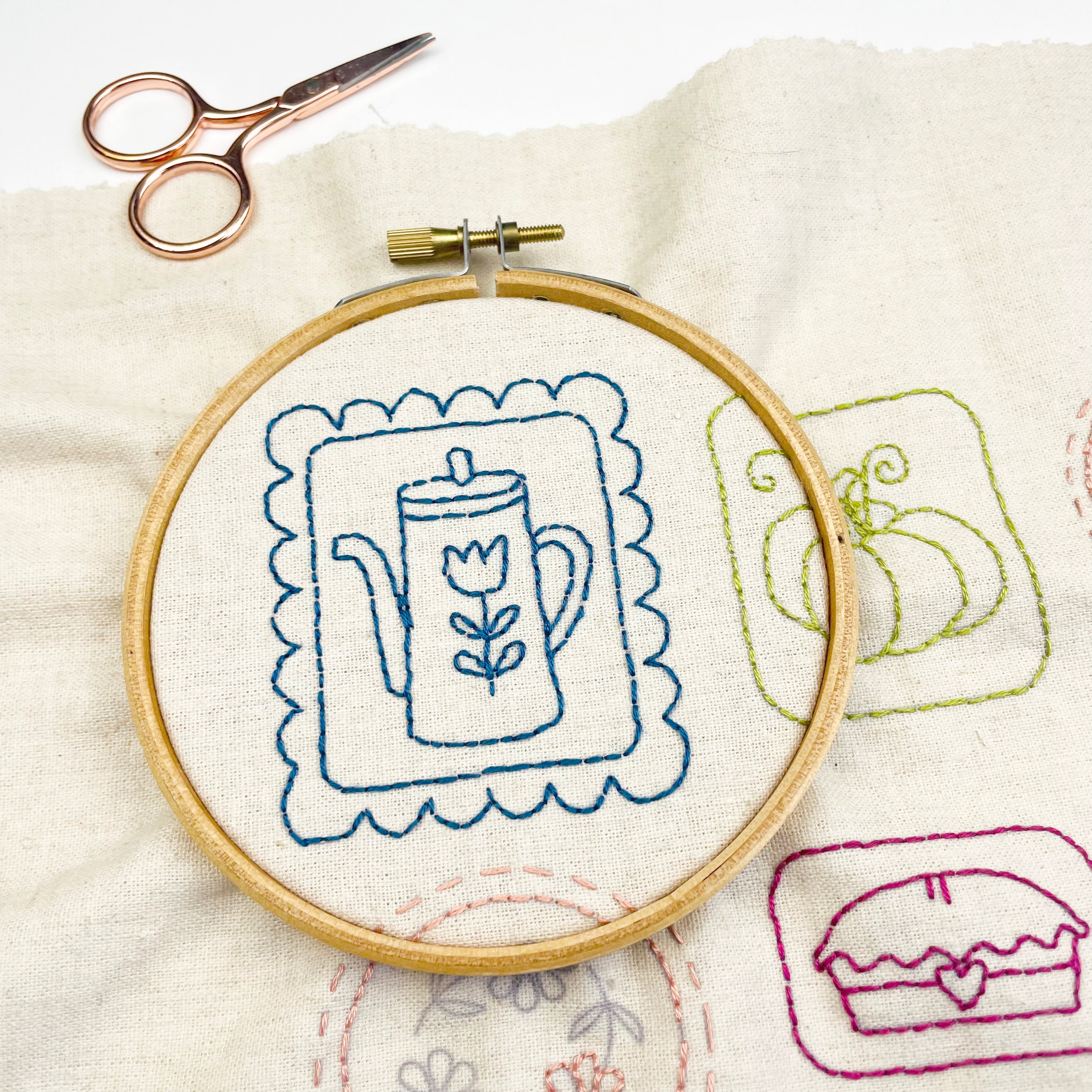 Embroidery hoop with blue embroidered teapot design on a beige fabric background