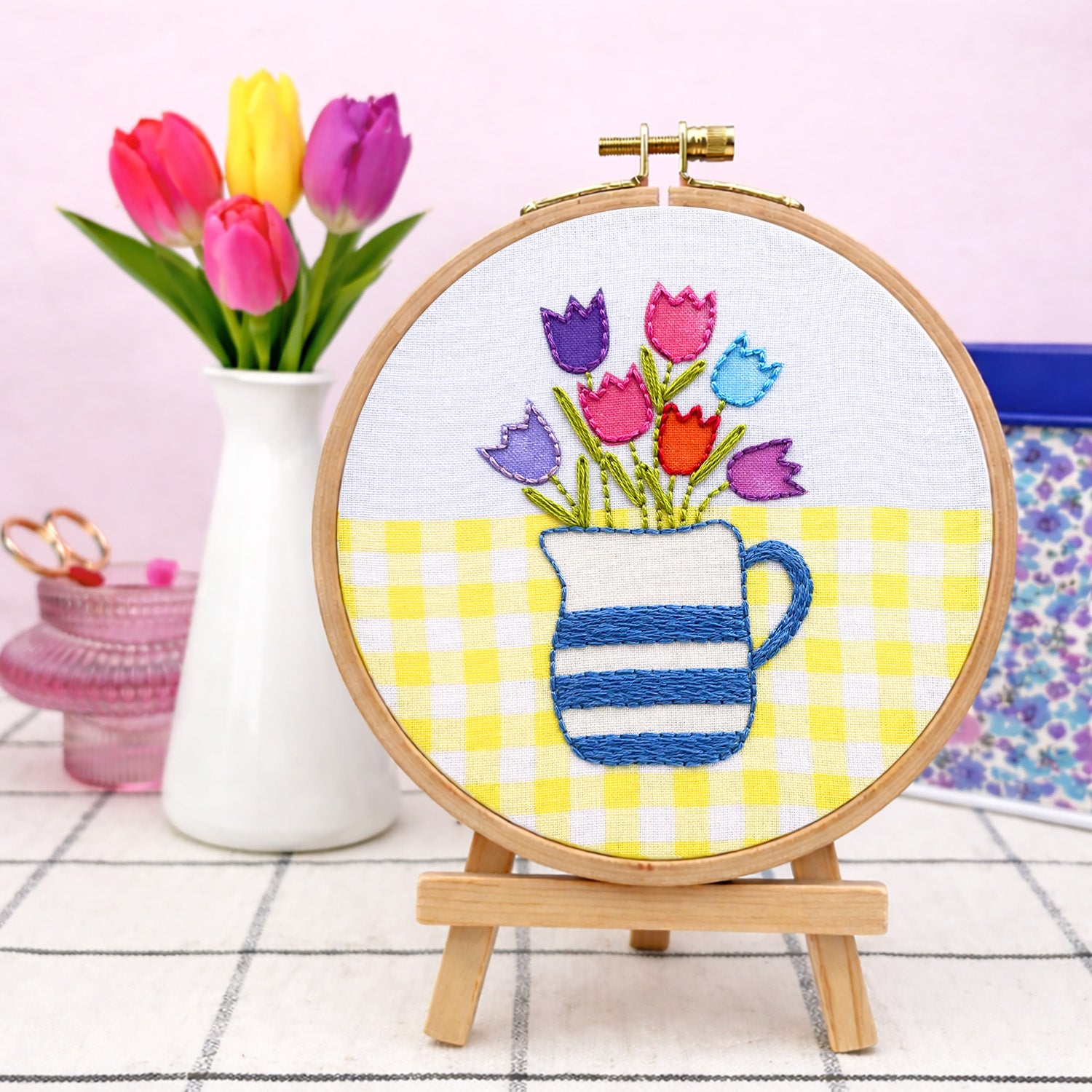 Embroidery hoop with floral design featuring tulips and a pitcher on a checkered background, with a vase of real tulips in the background.