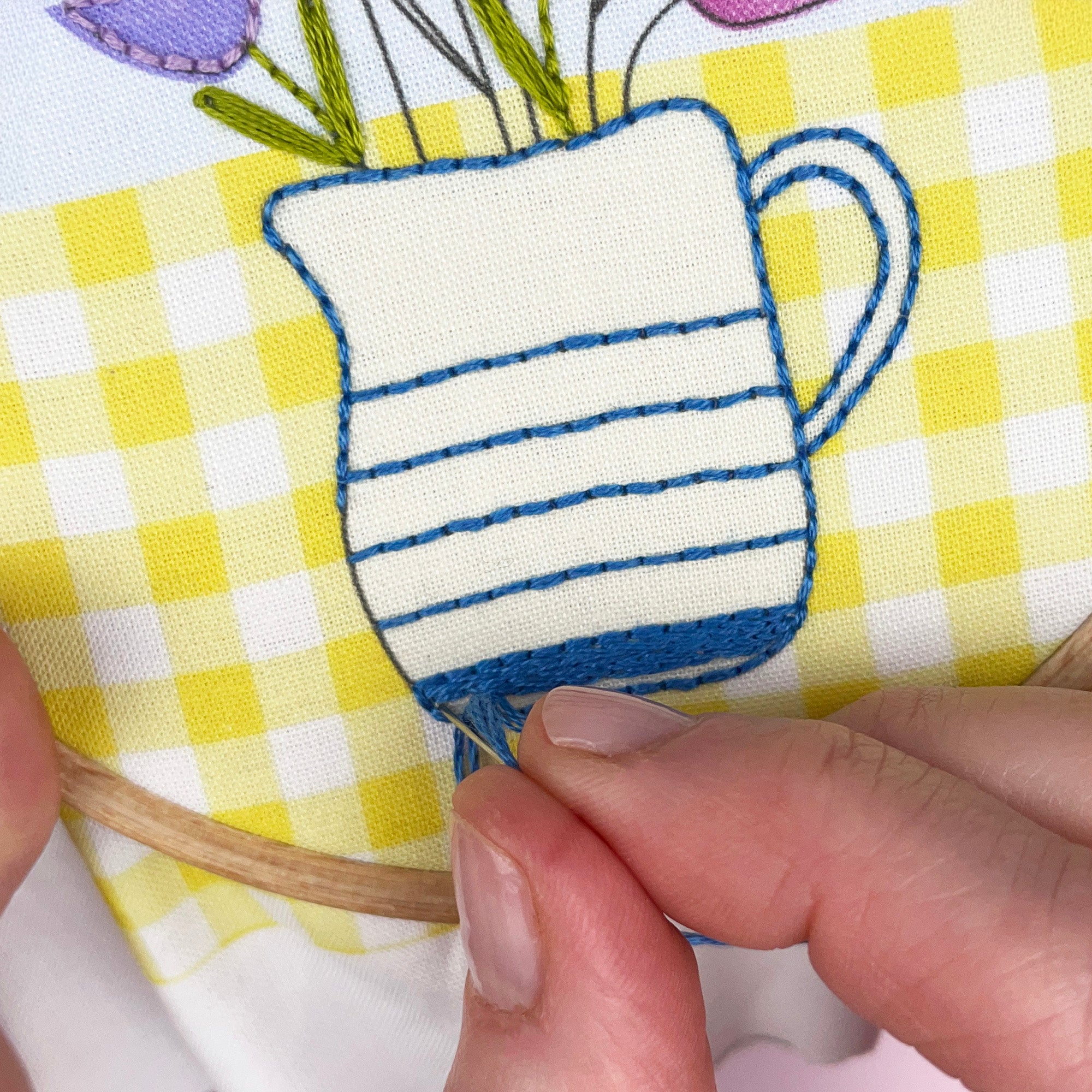 Embroidery of a blue and white striped mug with flowers on a yellow and white checkered fabric.
