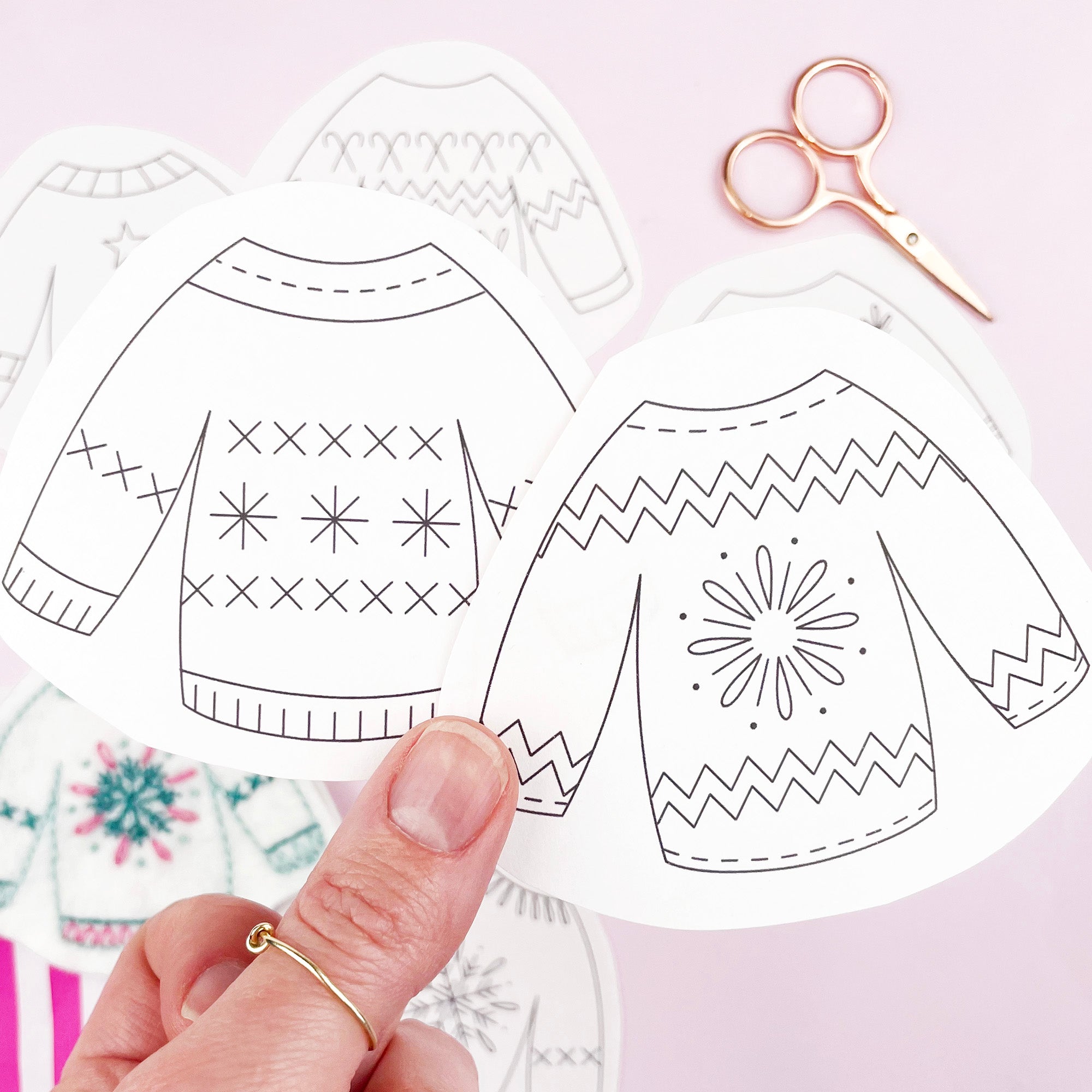 Two iron on embroidery patterns cut-out sweaters with patterns held by a hand against a light pink background.