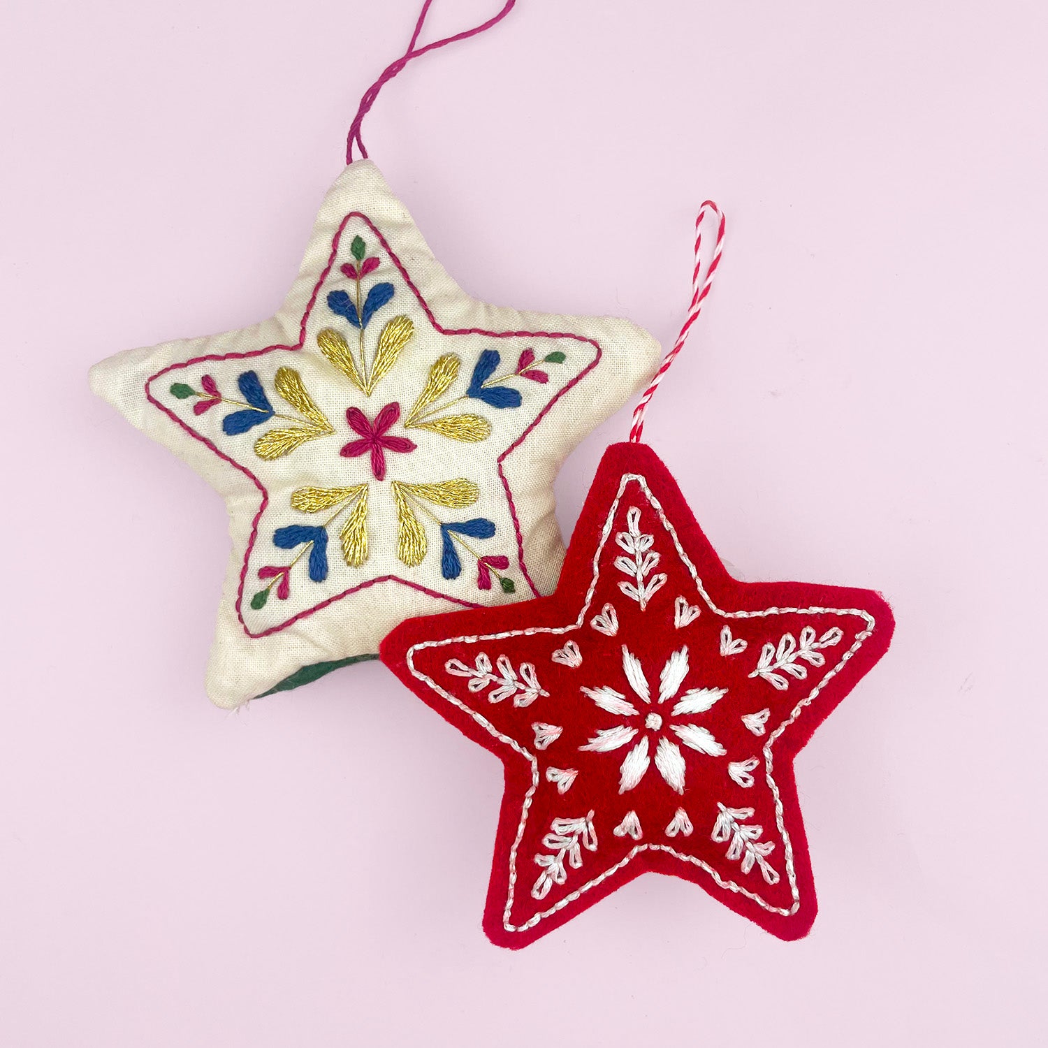 Two embroidered felt star Christmas ornaments hanging against a light pink background. One star is cream with multicolor floral embroidery, and the other is red with white floral embroidery.