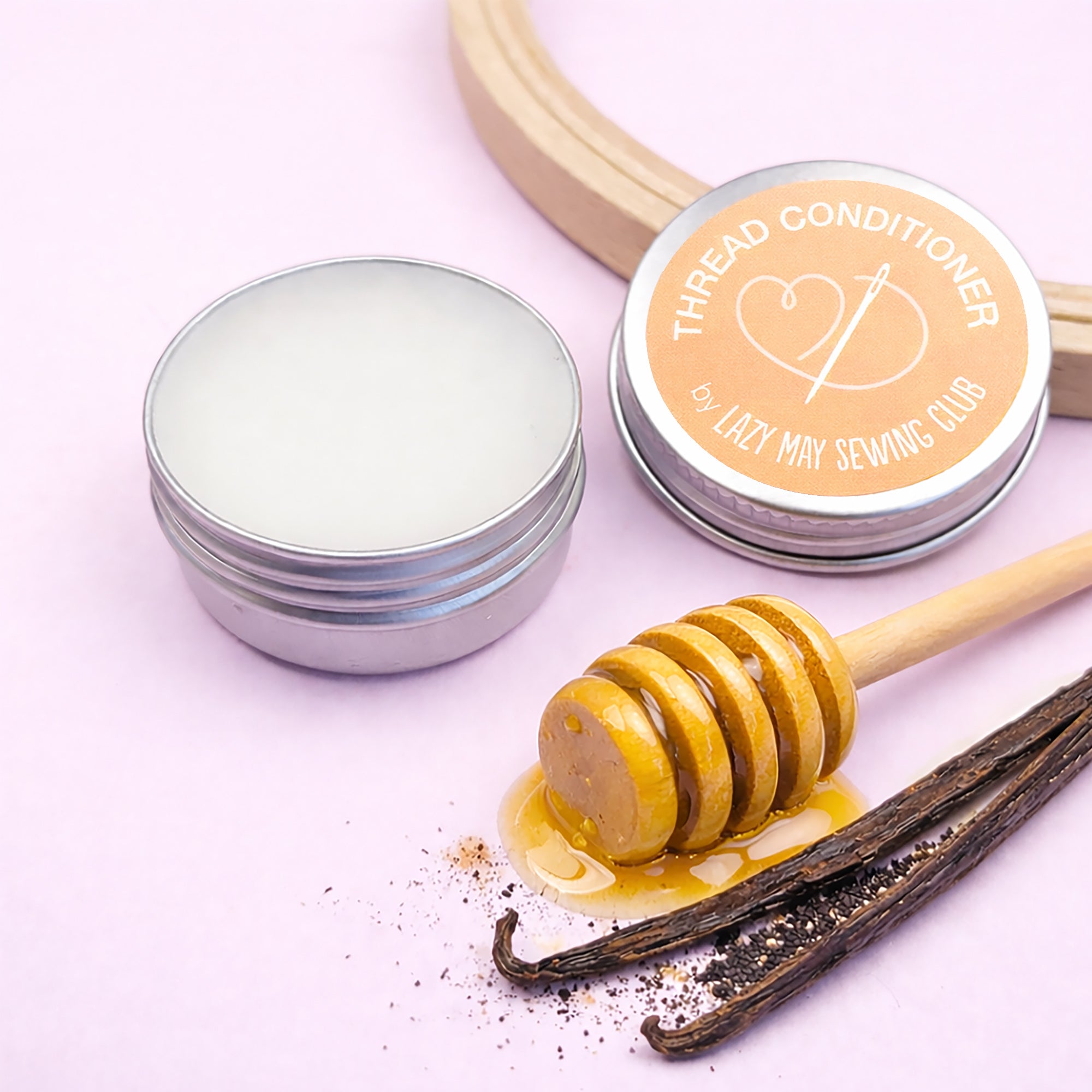 Tin of embroidery thread conditioner with lid, a honey dipper, and vanilla beans on a pink background.