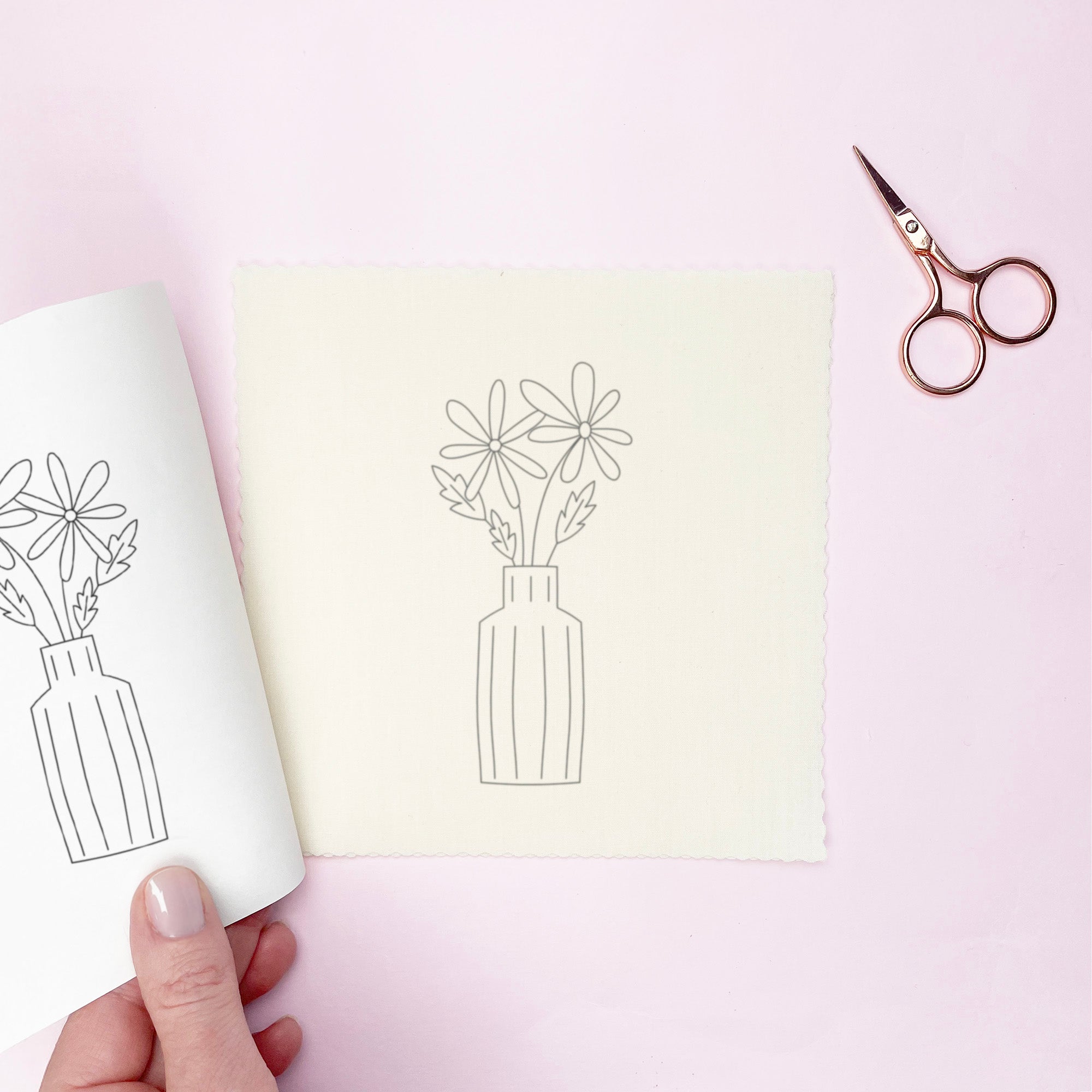 Hand holding a paper embroidery transfer with floral line drawing, scissors on a pink background