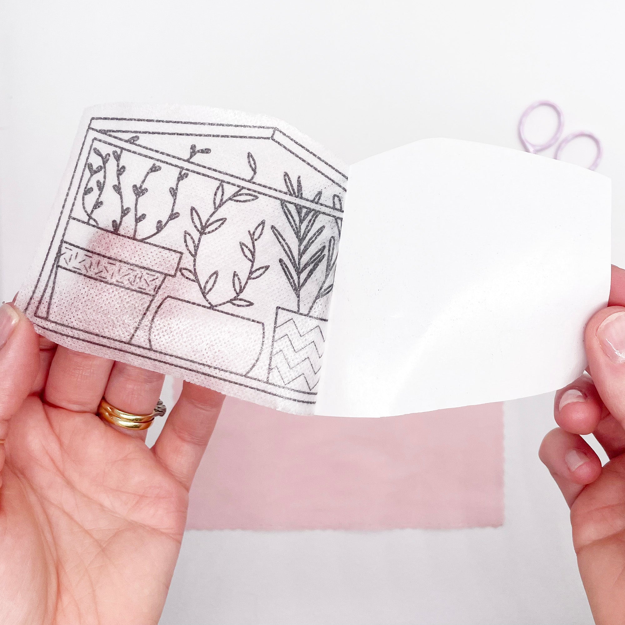 A hand peeling back the backing paper of a stick and stitch embroidery pattern featuring a greenhouse scene with potted plants.