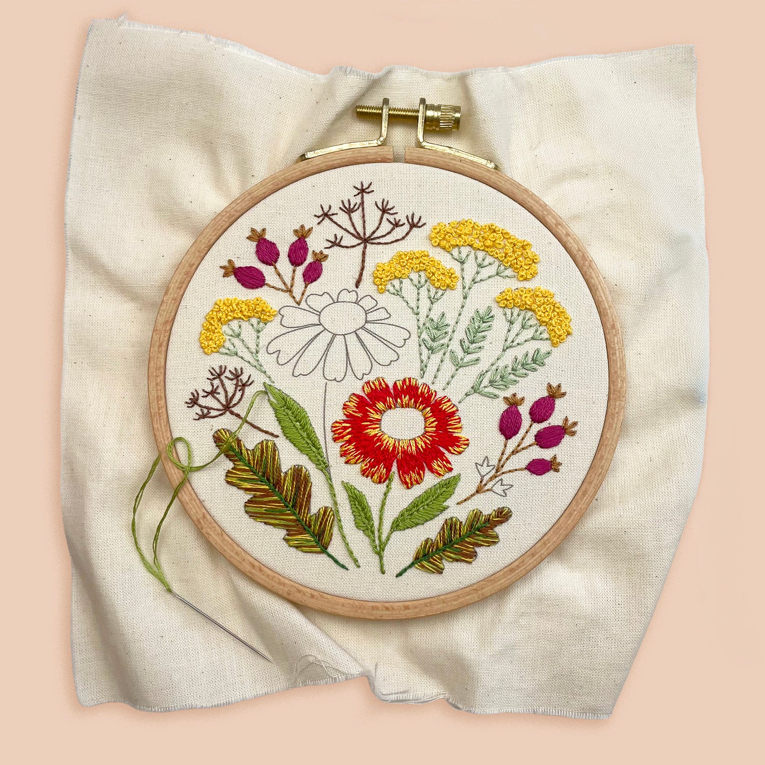 A circular embroidery hoop holding a floral needlepoint or embroidered design on a cream fabric background, photographed against a soft peach-pink backdrop.