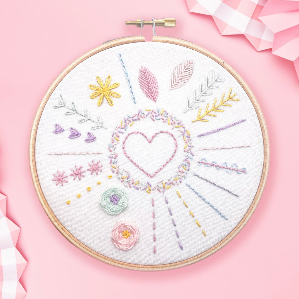 Embroidery sampler stitched in a hoop from a free PDF embroidery pattern. You can see 13 different embroidery stitches in pastel coloured threads on a pink background