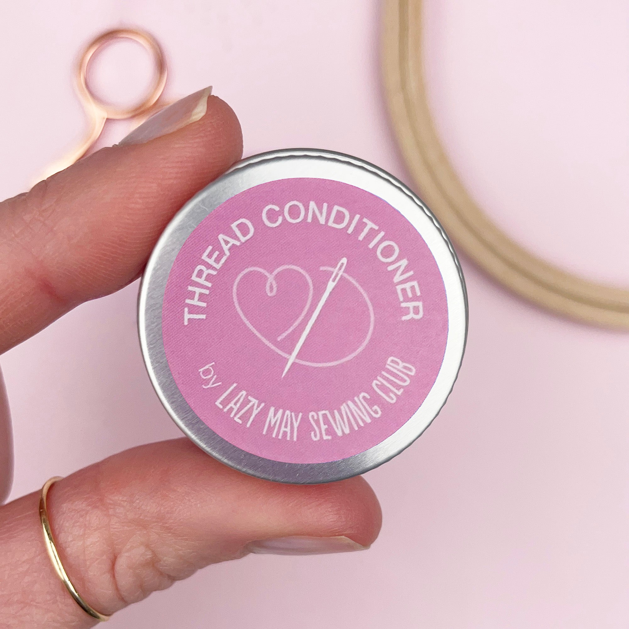 Hand holding a tin labeled 'Thread Conditioner' by Lazy May Sewing Club with sewing tools in the background.