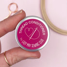 Hand holding a tin labeled 'Thread Conditioner by Lazy May Sewing Club' with sewing tools in the background.