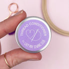 Hand holding a tin labeled 'Thread Conditioner' by Lazy May Sewing Club on a pink background.