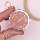 Hand holding a 'Thread Conditioner' container by Lazy May Sewing Club with sewing tools in the background.