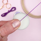 Person threading a needle with purple thread on a light pink background