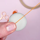 Close-up of a hand holding a spool of orange thread with scissors and a spool in the background on a light pink surface.