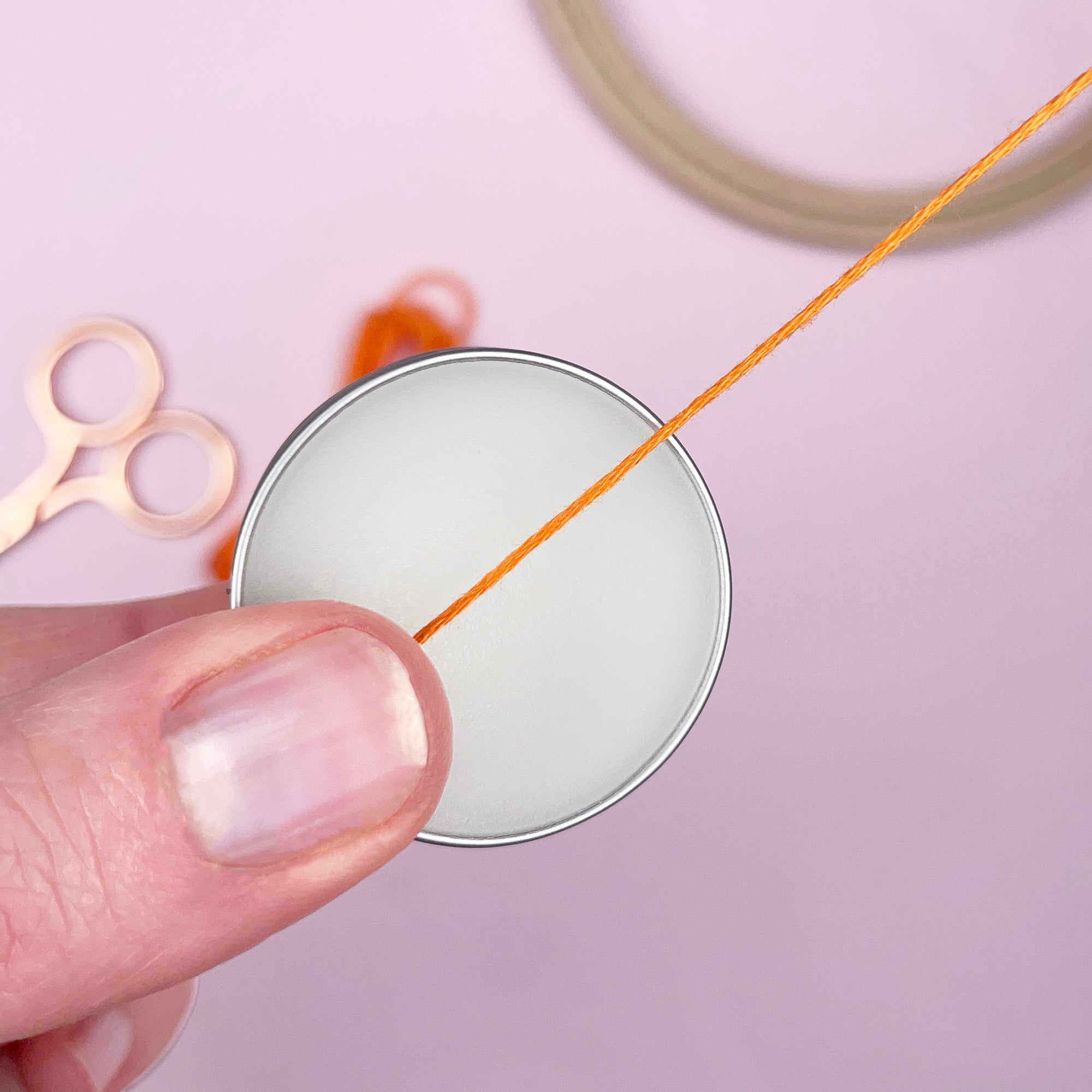 Close-up of a hand holding a spool of orange thread with scissors and a spool in the background on a light pink surface.
