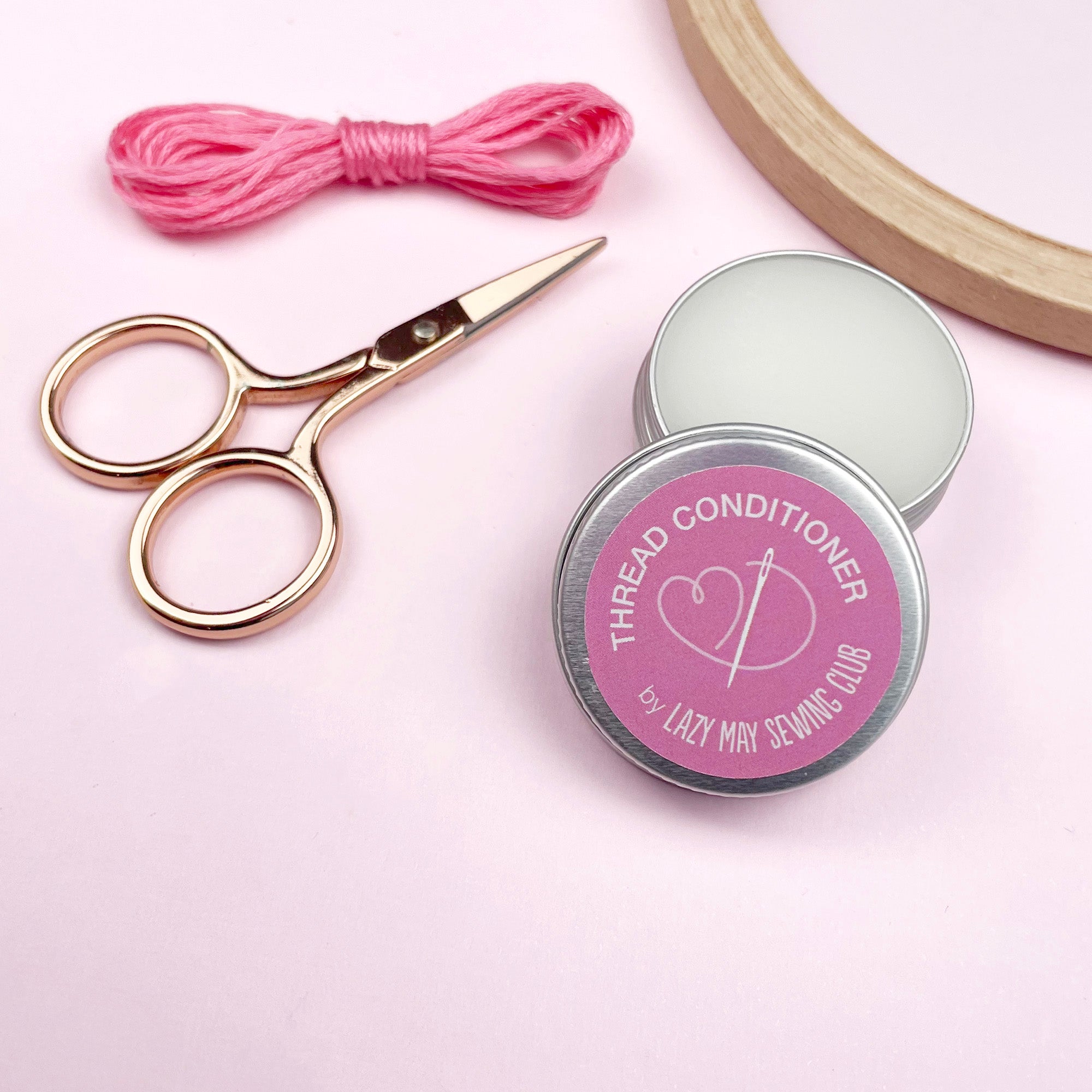 Thread conditioner tin with pink label, scissors, and pink thread on a light pink background