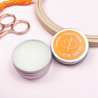 Thread conditioner container with a lid, scissors, and spool of thread on a light pink background