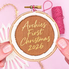 A circular wooden embroidery hoop ornament held by two hands against a pink background. The center wood panel is embroidered with gold script reading 'Archies First Christmas 2026', surrounded by white stitched border and a small red-and-white striped hanging loop.