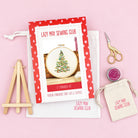 A flat-lay of a Lazy May Sewing Club 5" embroidery kit on a light pink background, including an embroidery instruction booklet, a small wooden easel, a white canvas bag, gold-handled scissors, a small tin of thread, and a matching drawstring pouch.