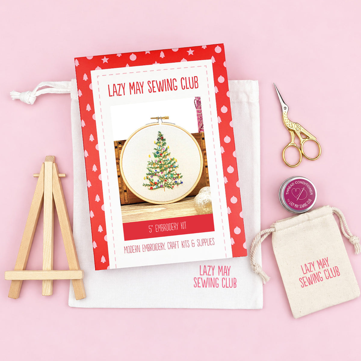 A flat-lay of a Lazy May Sewing Club 5" embroidery kit on a light pink background, including an embroidery instruction booklet, a small wooden easel, a white canvas bag, gold-handled scissors, a small tin of thread, and a matching drawstring pouch.