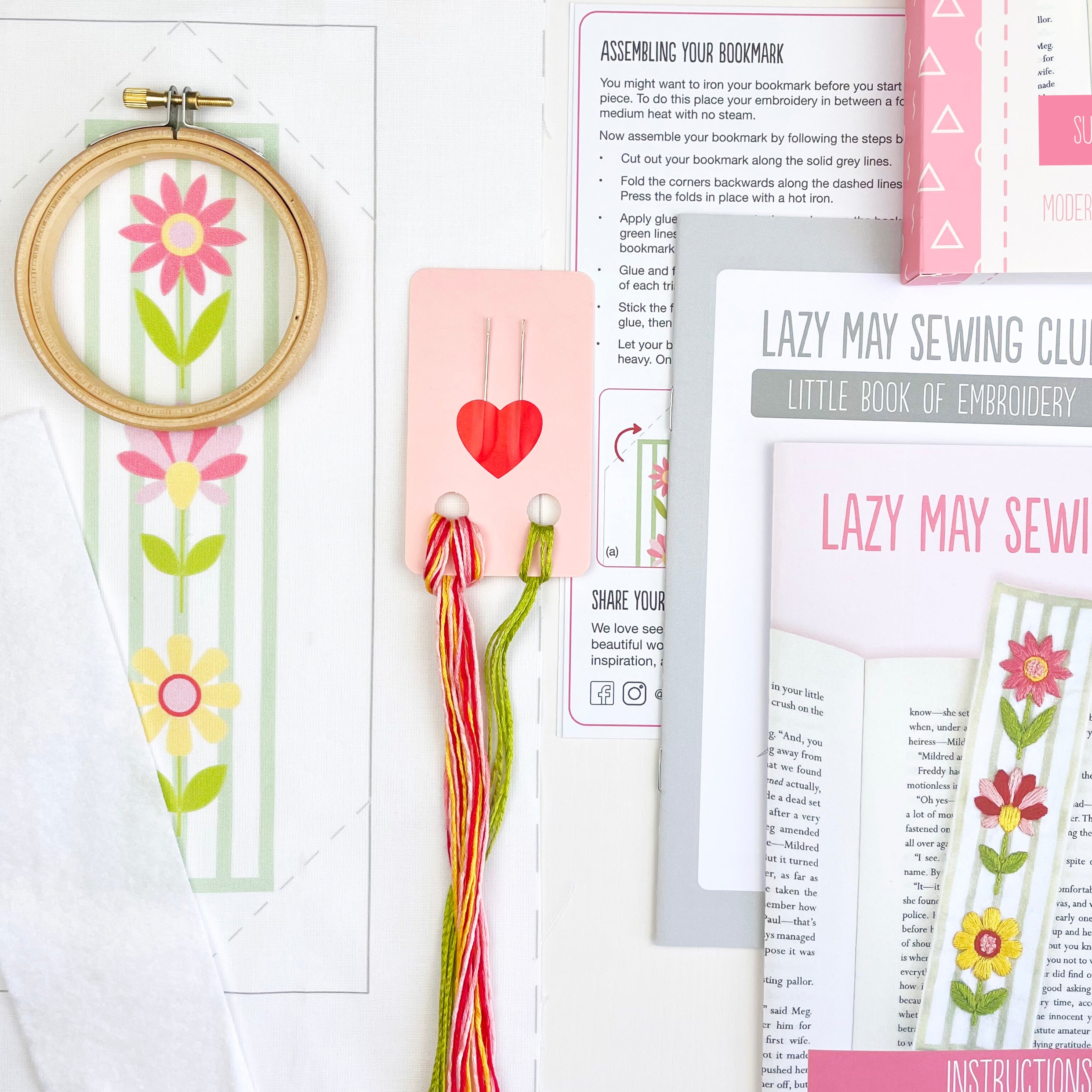 Content of a kit to embroider your own bookmark. Embroidery hoop, fabric printed with floral design, needles, thread and instructions, and 'Lazy May Sewing Club' beginners embroidery booklet on a white background
