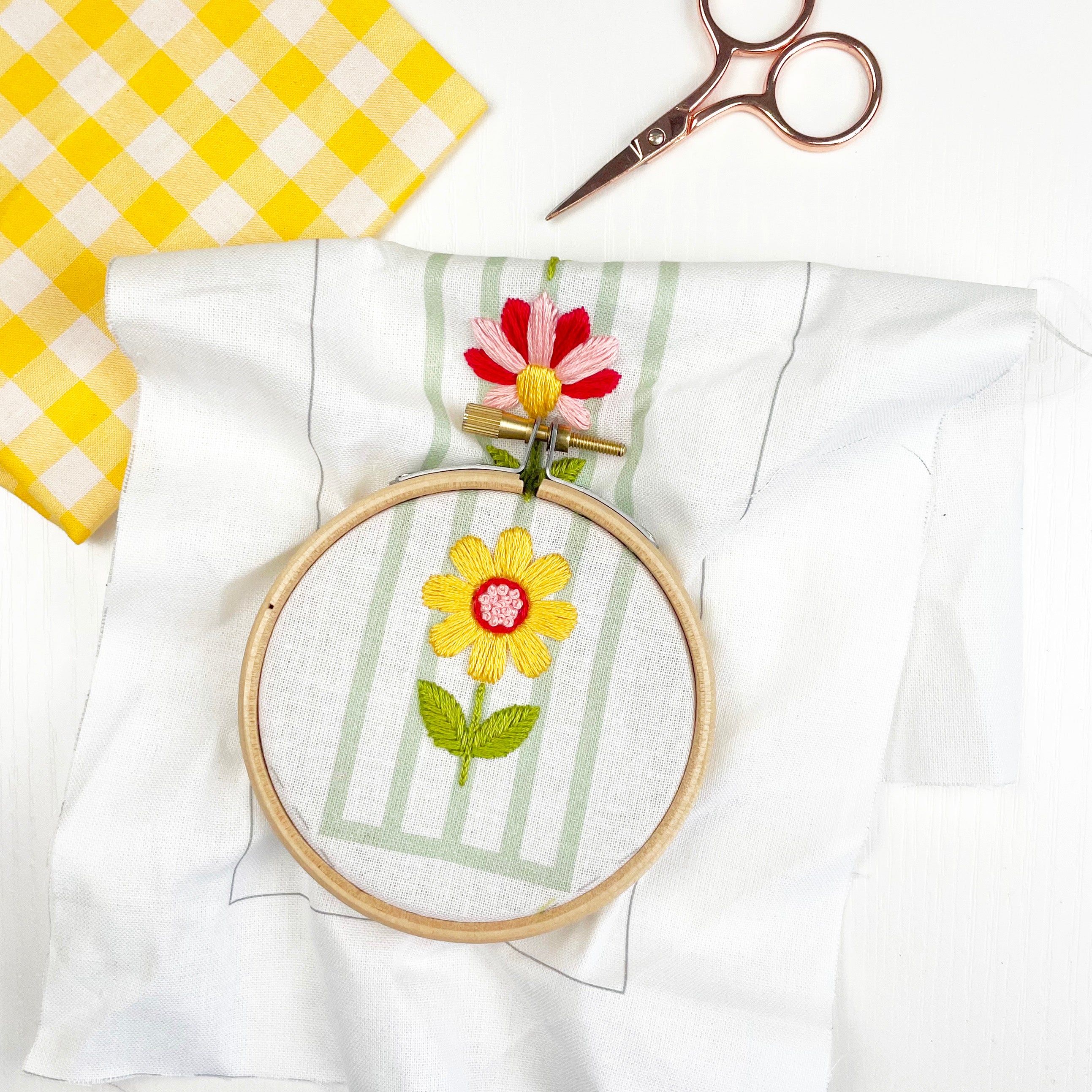 Embroidery hoop with floral design on a white fabric background. In-progress stitching of a flower using a 3" embroidery hoop. Easy embroidery bookmark project using pre-printed fabric and thread.