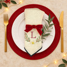 A festive table setting featuring a cream-colored napkin with a red velvet bow, embroidered monogram 'L', and holiday wreath details, styled on a white plate with a red charger placemat, gold cutlery, and green leaves.