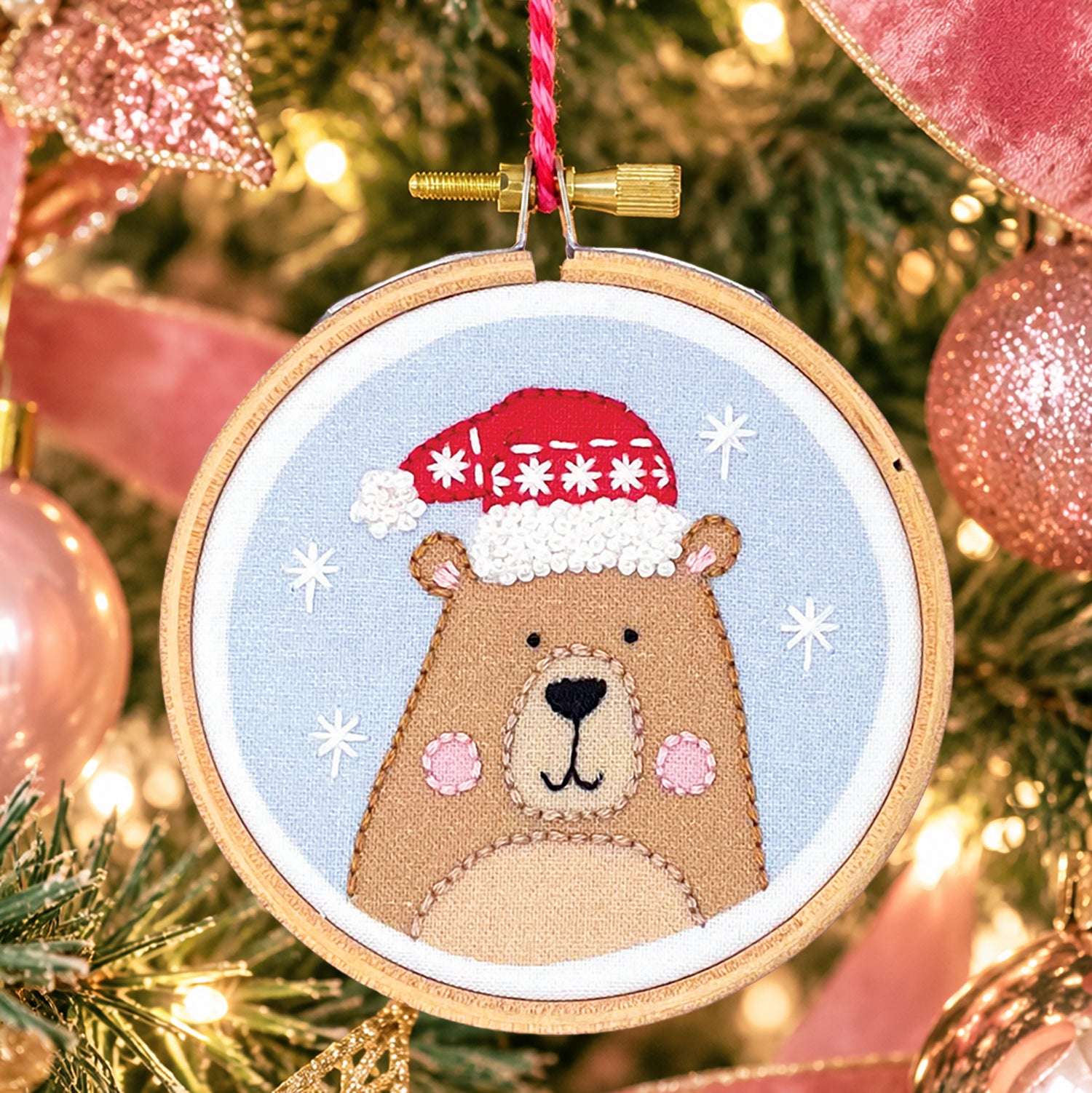A circular embroidery hoop ornament featuring a stitched brown bear wearing a red Santa hat on a light blue background, hanging in front of a softly blurred Christmas tree with pink ornaments and warm lights.