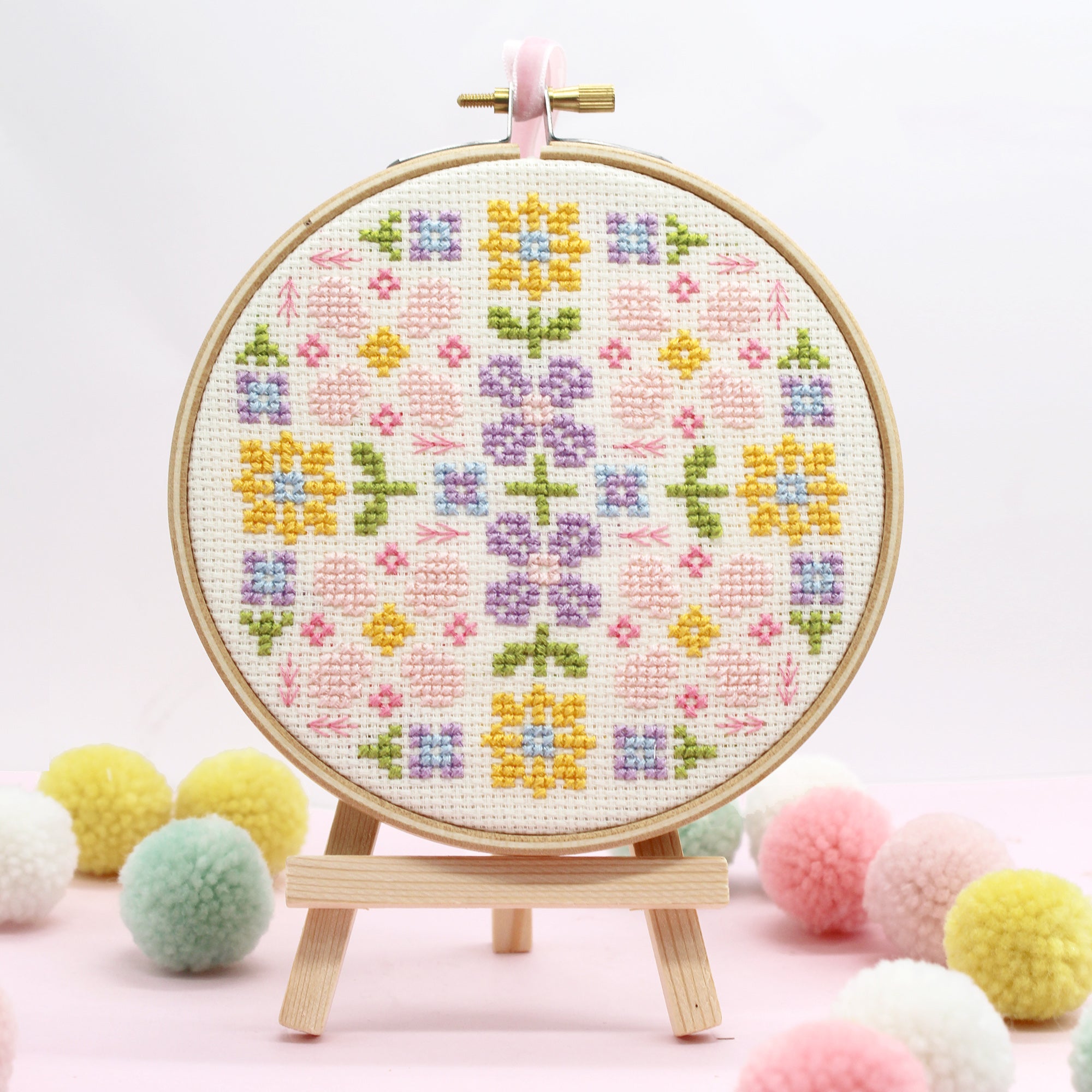 Close-up of completed floral cross-stitch design in an embroidery hoop, featuring pastel flowers in pink, yellow, blue, and green. Displayed on a small wooden easel with soft pom-poms in the background