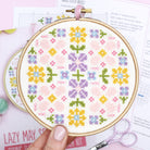 A close-up of a completed floral cross-stitch design displayed in an embroidery hoop. The pattern features a vibrant bouquet of spring flowers in shades of pink, yellow, blue, and green, showcasing intricate stitching details.