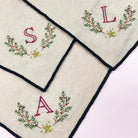 Three square cream linen napkins with black borders, each decorated with a green and red embroidered wreath, a gold star, and a monogram letter (S, L, and A respectively) on a light pink background.