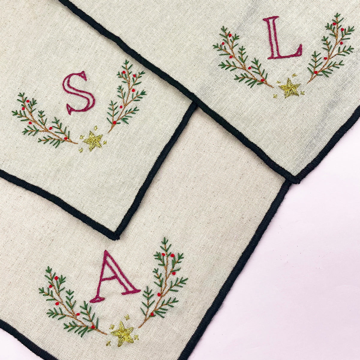 Three square cream linen napkins with black borders, each decorated with a green and red embroidered wreath, a gold star, and a monogram letter (S, L, and A respectively) on a light pink background.