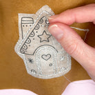 Hand sewing a decorative patch with a house design onto a brown felt fabric background.