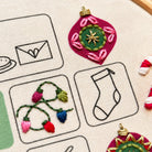 Embroidery advent calendar. Close-up of a hand-embroidered canvas patch featuring a magenta Christmas ornament with a green center and gold star, alongside other embroidered motifs on a beige fabric background.