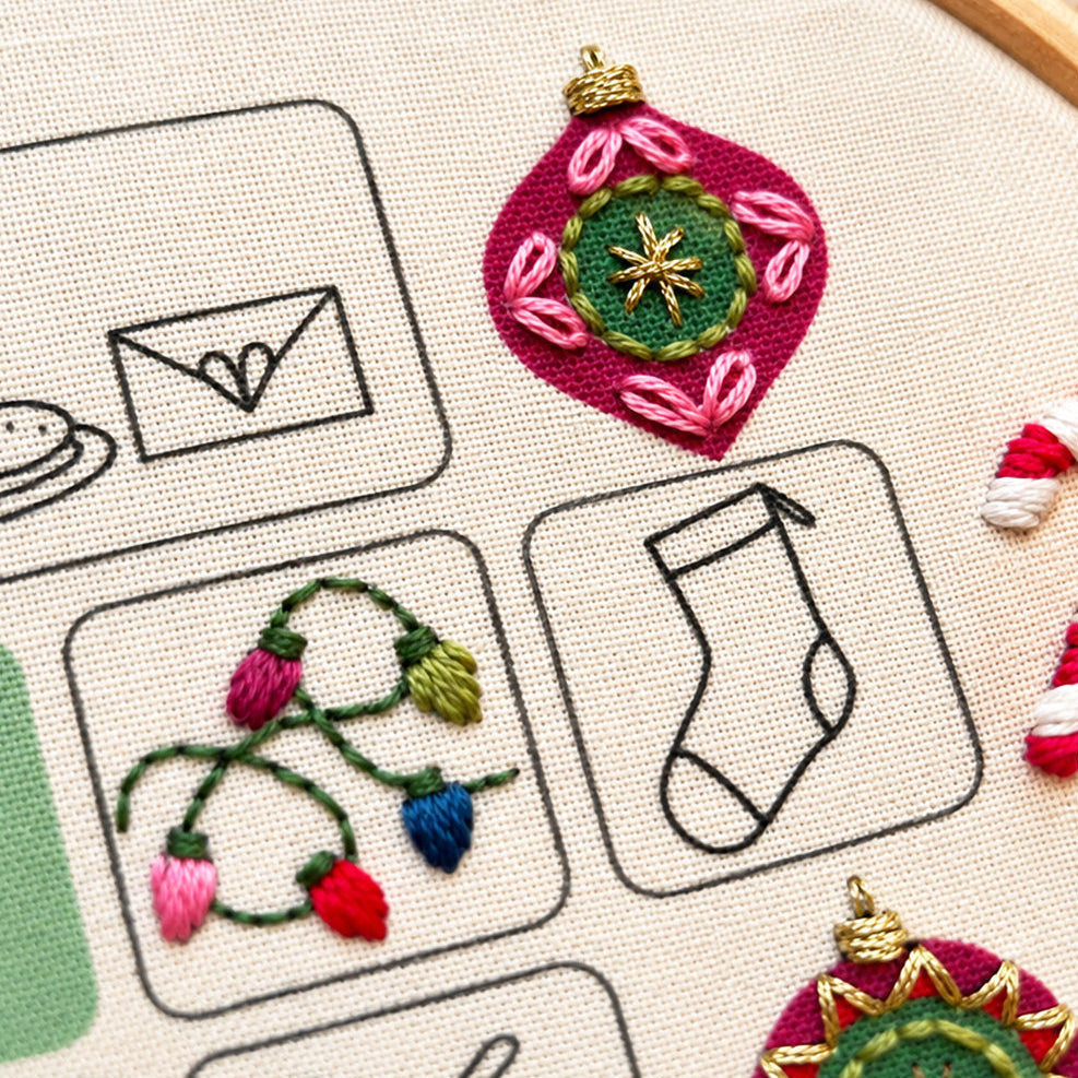 Embroidery advent calendar. Close-up of a hand-embroidered canvas patch featuring a magenta Christmas ornament with a green center and gold star, alongside other embroidered motifs on a beige fabric background.