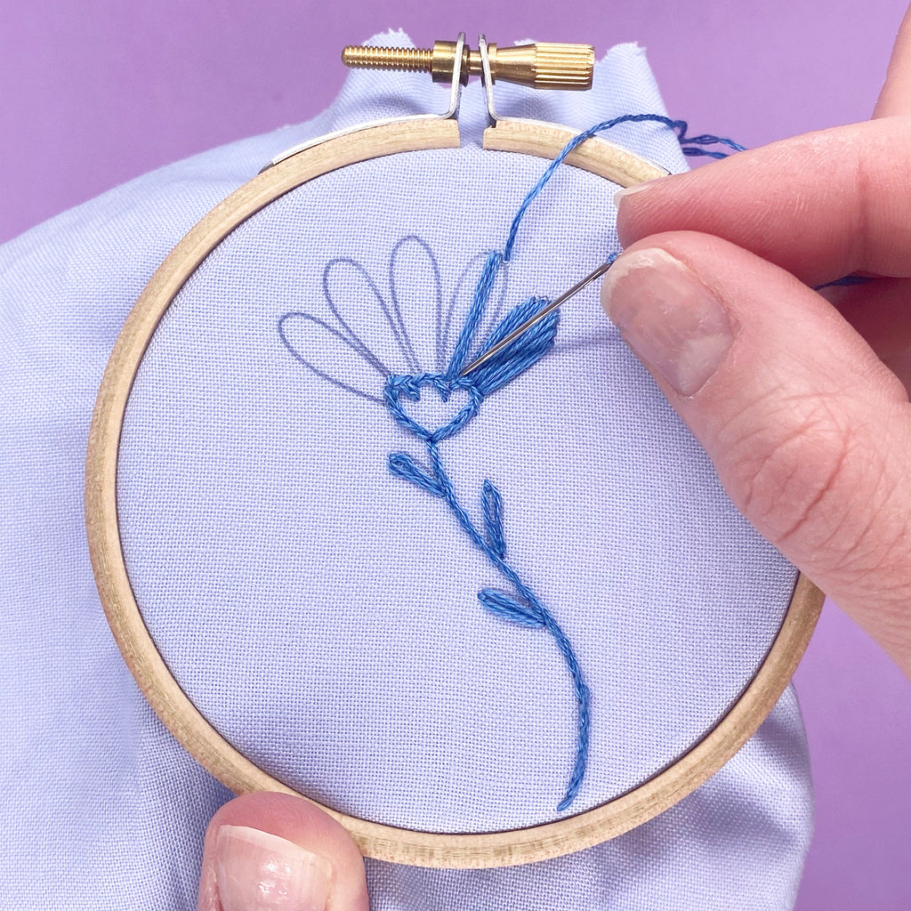 blue flower being embroidery by hand with blue embroidery thread. the embroidery is in a wooden hoop and has a modern flower design from an iron on embroidery patterns