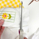 Hand holding scissors cutting a piece of fabric with a floral design on a checkered background. Part of a sew your own floral embroidery bookmark kit for beginners.