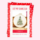 A Christmas-themed embroidery kit package from Lazy May Sewing Club, featuring a red and white patterned border and a central photo of a completed embroidered Christmas tree in an embroidery hoop.