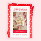 A boxed embroidery kit package titled 'Lazy May Sewing Club' with a red-and-white polka-dot border and a central photo of a star-shaped embroidered ornament. The package is photographed on a pale pink background.