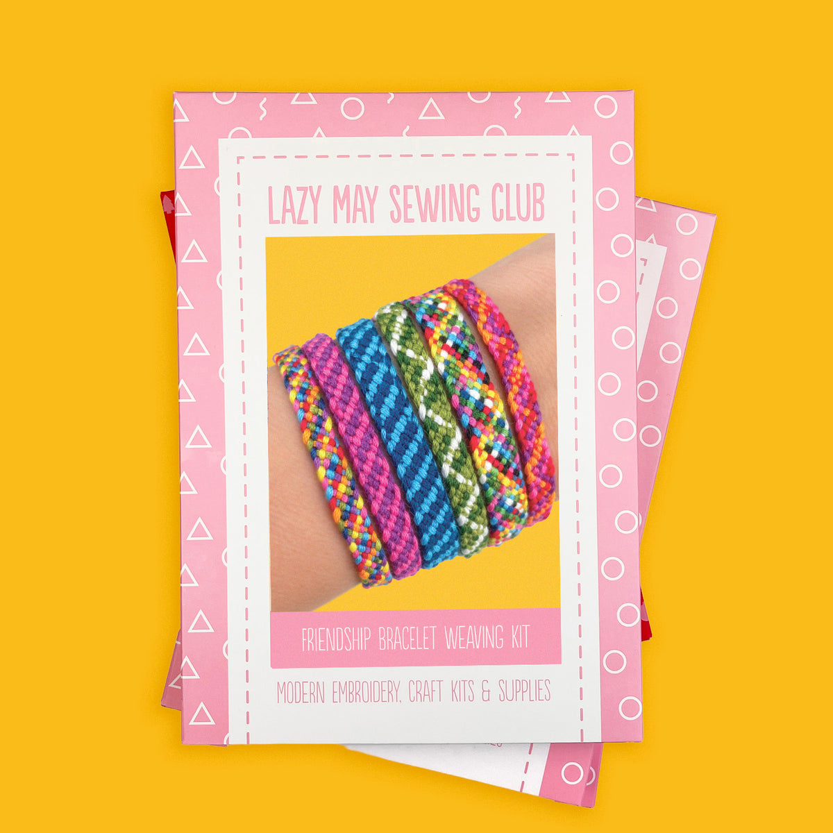 A pink and white boxed kit titled 'Lazy May Sewing Club Friendship Bracelet Weaving Kit' is shown on a bright yellow background, with a photo of colorful woven bracelets on a wrist printed on the cover. 