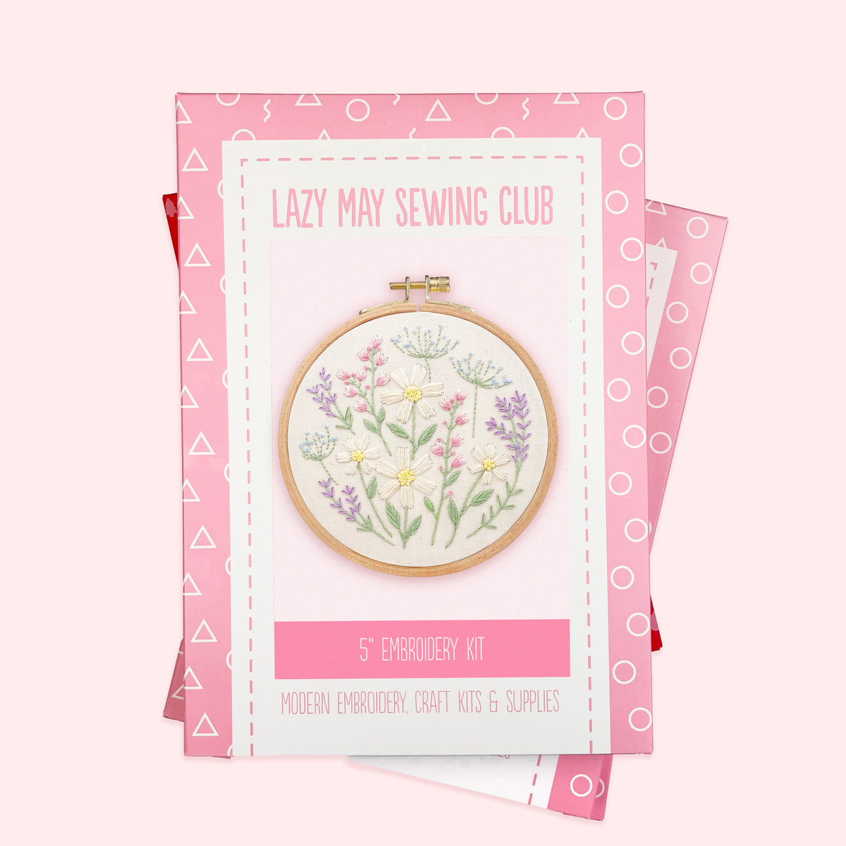 A 5" embroidery kit by Lazy May Sewing Club, featuring a floral design in a wooden hoop, presented on pink patterned packaging against a pale pink background.