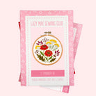 A pink and white embroidery kit box labeled 'Lazy May Sewing Club' with a floral embroidery design in the center, shown on a pale pink background with additional matching boxes partially visible behind it.