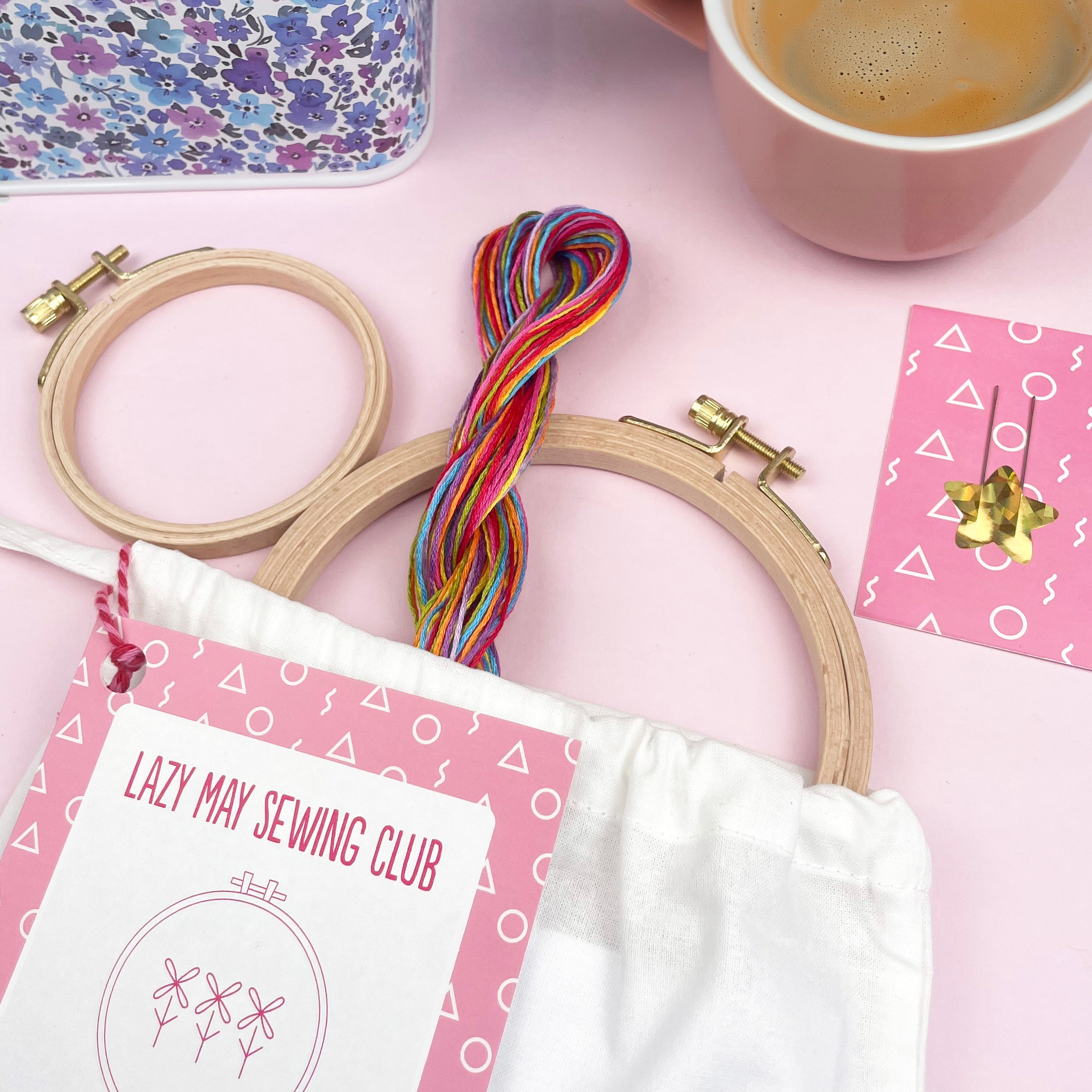 Colorful embroidery threads and hoops on a pink background with a coffee cup and sewing club materials
